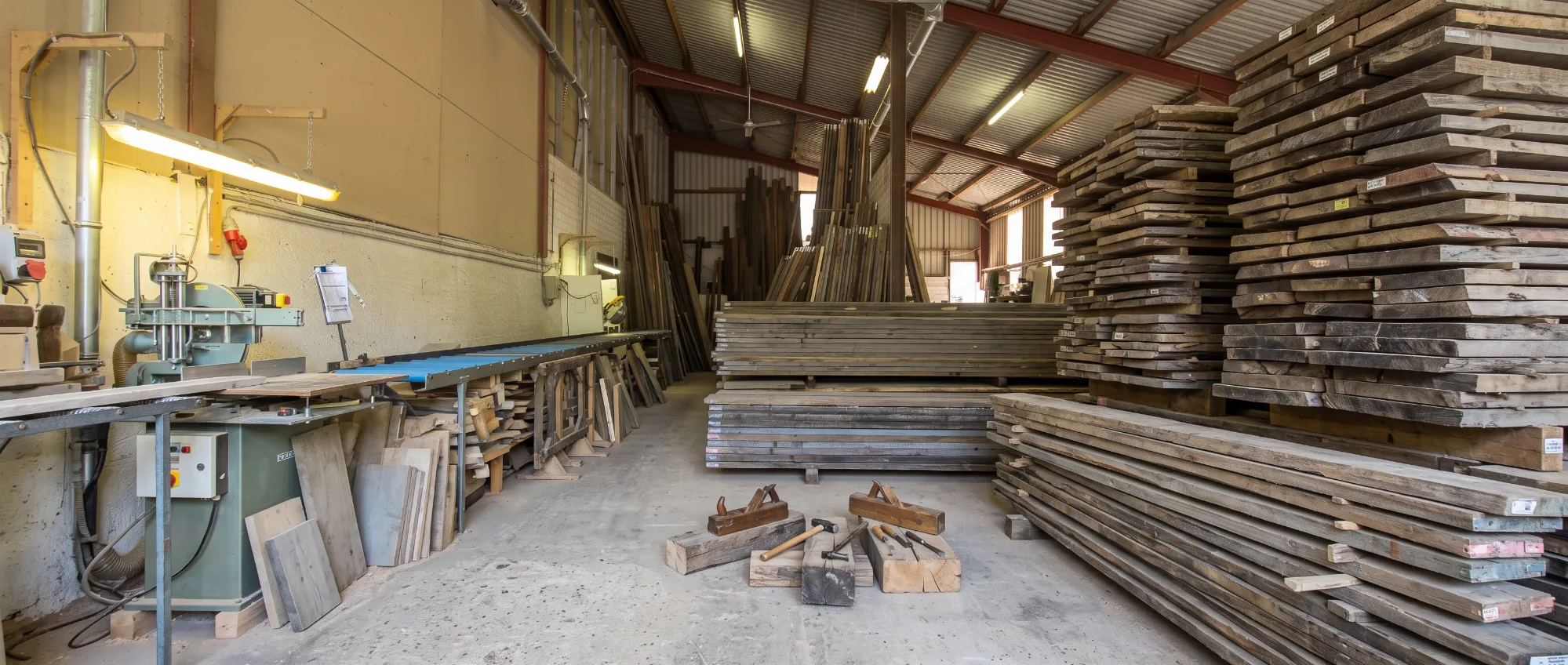Ein Blick in die Produktionshalle der Tischlerei Brand: Große Stapel aus historischem Bauholz und professionelle Holzbearbeitungsmaschinen.